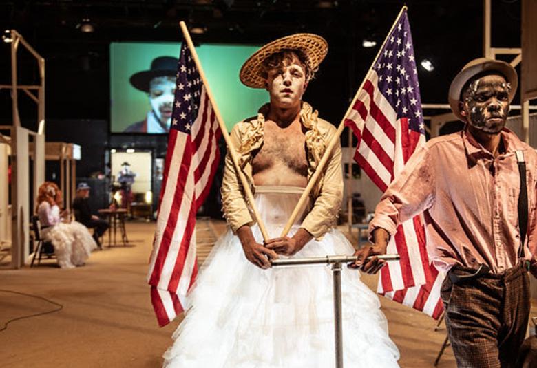 Kuvassa on edessä vasemmalla tyttö, jolla on käsissään kaksi isoa Amerikan lippua. Tytöllä on päällään kullan värinen jakku ja sen alta näkyy rintakarvoja sekä valkoinen röyhelöhame, joka on pitkä. Vieressä on mustaihoinen nuorimies, jolla on vaalean ruskea paita ja tumman ruskeat housut sekä päässään musta hattu. Taustalla on telineitä ja seinällä on iso näyttö ja siinä mustan miehen kasvot ja hänellä on musta hattu.