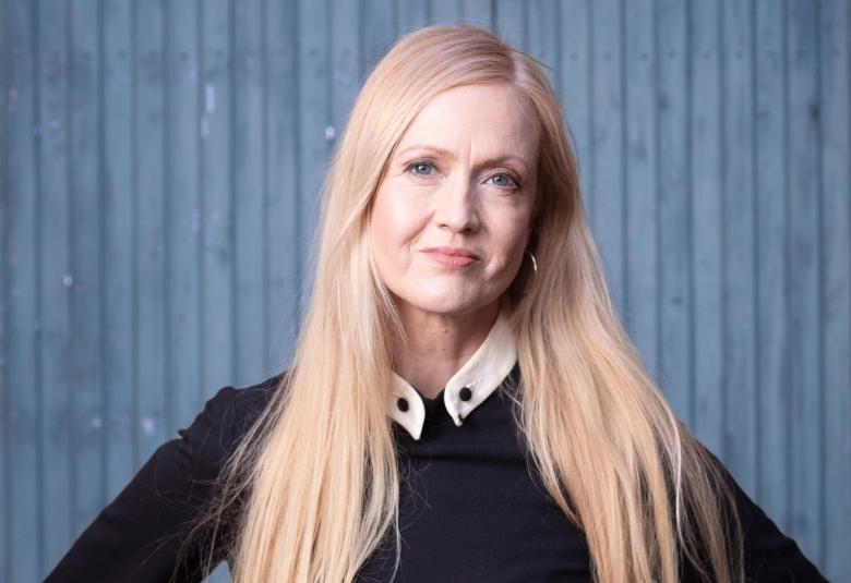Kuvassa on Eveliina Talvitie mustassa puserossa puolivartalokuvassa ja hänellä on kädet lantiolla. Hänellä on pitkät vaaleat hiukset. Tausta on harmaan sininen.