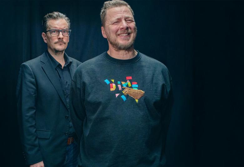 Kuvassa on etualalla oikealla Jaakko Saariluoma tumman sinisessä college-paidassa, jossa on kuvio.  Takana on André Wickström mustassa takissa. Tausta on tumma.