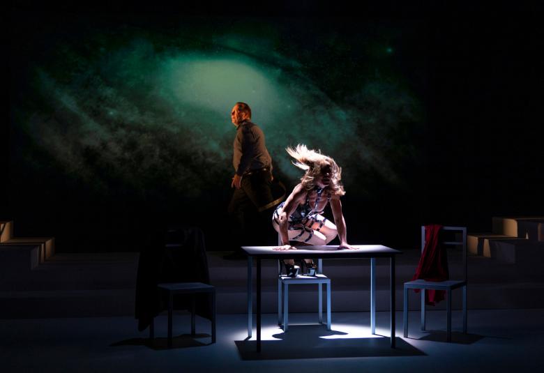 Kuvassa on tummalla näyttämöllä valaistuna edessä pöydän päällä nainen polvillaan ja hänellä on valkoiset ihonmyötäiset vaatteet.  Hänen hiuksensä hulmuavat tuuletuksessa. Taustalla näkyy mieshenkilö seisomassa.