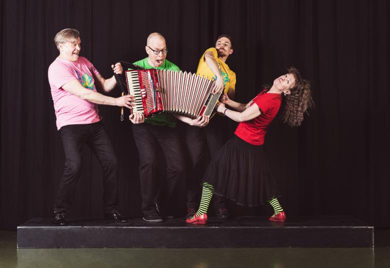 Kuvassa ovat hieman korotetulla lavalla Loiskiksen neljä jäsentä ja he kiskovat keskellään olevaa harmonikkaa. Tausta on tumma. 