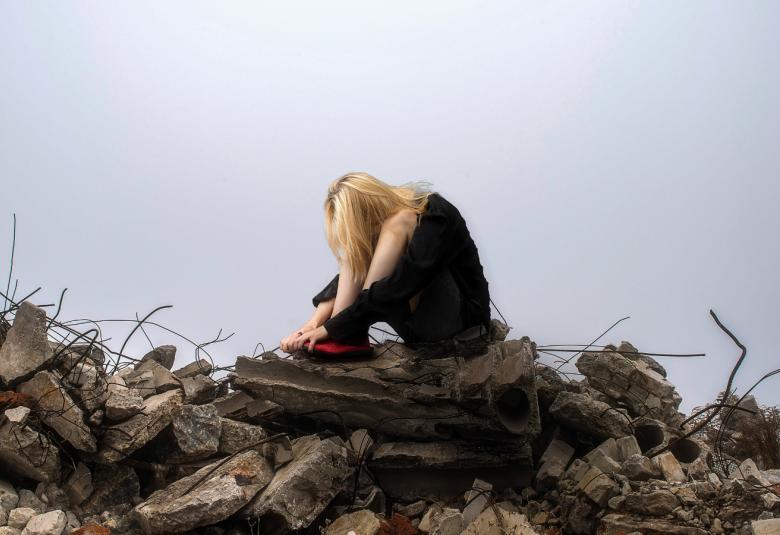 Kuvassa on kivisen ja betonisen kasan päällä, jossa näkyy raudoituksia, nuori nainen kyyristyneenä kädet polvien ympärillä.  Taustalla on sinistä taivasta.