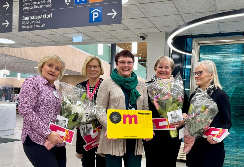 Kuvassa on viisi naista seisomassa ja heillä on kukkia kädessään sekä keskimmäisellä iso museokortti.  Taustalla näkyy ylhäällä kylttejä, joissa on nuolia ja ohjeita, minne mennä.