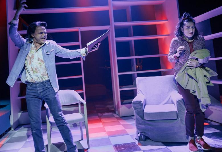 Kuvassa on vasemmalla nuori mies farkkupuvussa ja vaaleassa paidassa. Hän tanssii ja hänellä on oikea käsi pystyssä sekä vasemmassa kädessään papereita .  Oikealla on nainen pitkissä housuissa ja takki kädessään. Takana on kaksi nojatuolia. Taustalla on vaaleita hyllyjä. Kuvan sävy on sininen. 