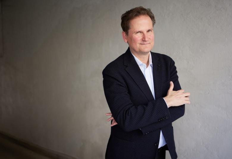 Kuvassa on Olli Mustonen mustassa takissa ja kädet ristikkäin rinnalla.  Taustalla on tumman harmaata käytävää.