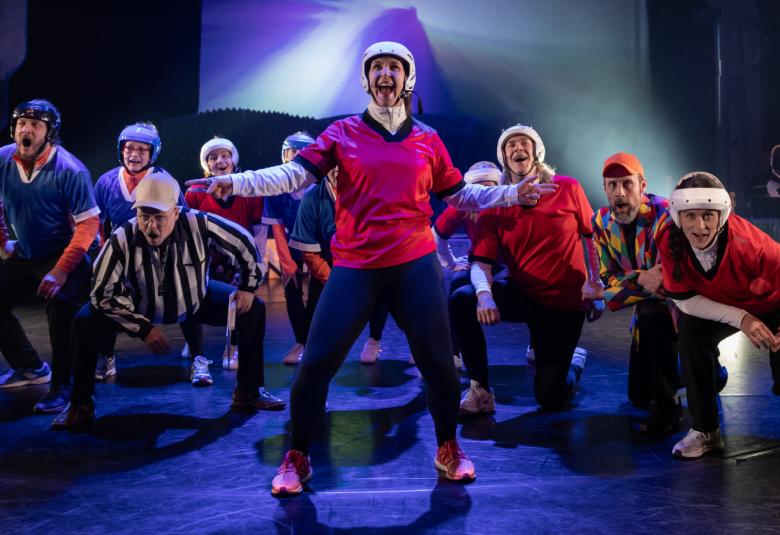 Kuvassa on taustalla kaarella näyttelijät punaisissa, valkoisissa tai sinisissä pelipaidoissa ja kypärissä. Keskellä on yksi miesnäyttelijä seisomassa punaisessa paidassa ja tummissa housuissa. Näyttämön sävy on tumma.