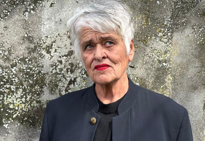 Kuvassa on Pirkko Saisio puolivartalokuvassa seisomassa mustassa jakussa.Taustall on vaalean harmaata sammaloitunutta kalliota. 