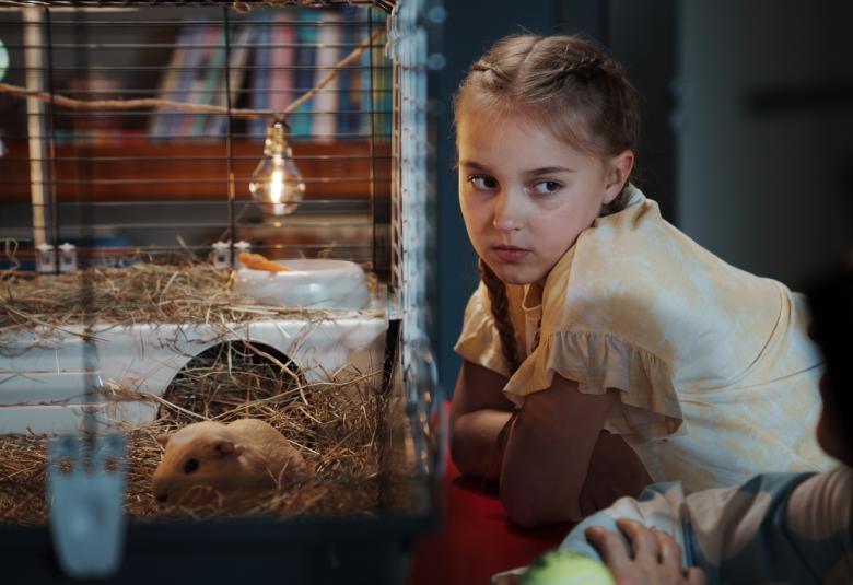 Kuvassa on Siiri Virtanen vaaleassa puserossa istumassa huoneessa ja katsoo pientä marsua häkissä. Taustalla on roikkuvia lamppuja.