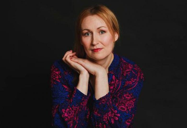 Kuvassa on Tiina Raevaara istumassa puolivartalokuvassa. Tausta on tumma. Hänellä on punaiset hiukset takana ja kämmenet yhdessä kasvojen alapuolella. Hänellä on sini-punakirjava pusero.