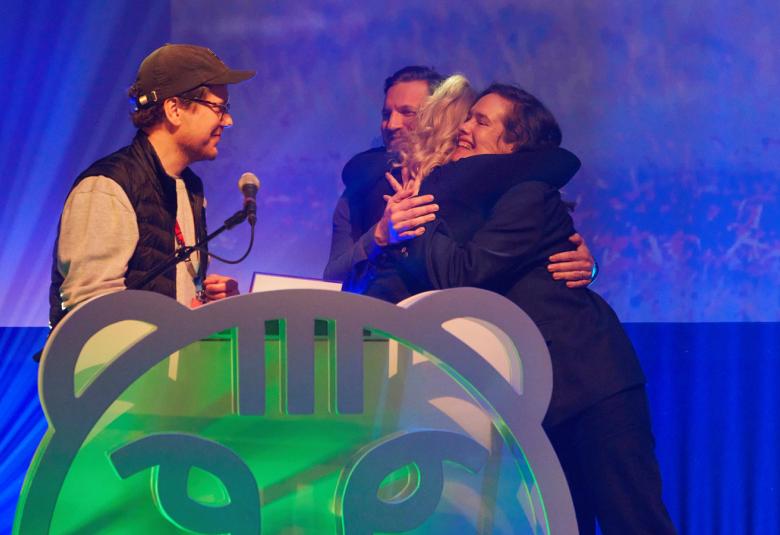 Kuvassa on vasemmalla mies lippalakissa mikrofoni kädessään ja hänen edessään mies ja nainen lasinen palkinto kädessään.  Edessä on puolikas, vihreä muovinen kissanpää. Tausta on sähkönsininen.