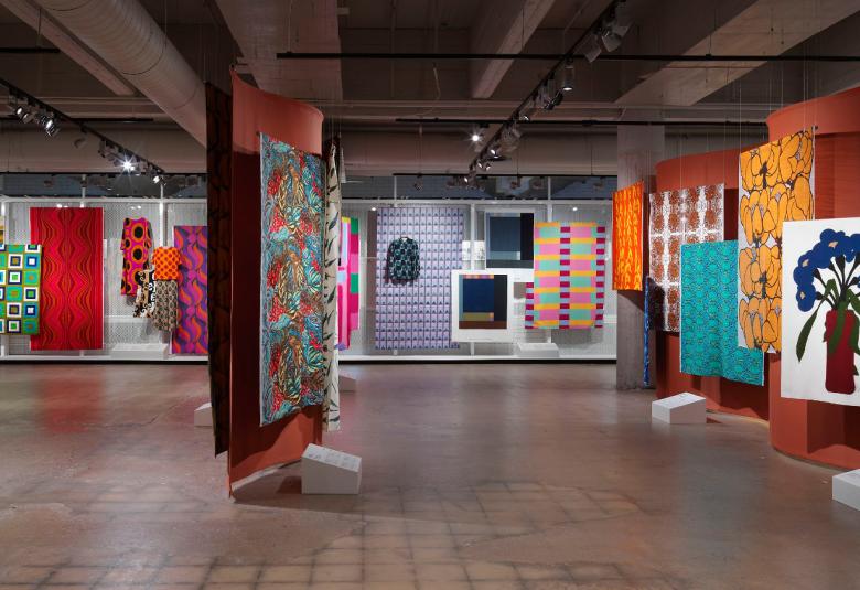 Kuvassa on näyttelytila, joka on jaettu seinämällä keskioikealta. Seinillä on värikkäitä tekstiilikankaita.