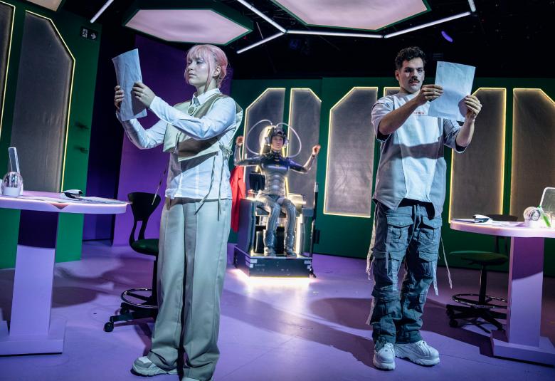 Kuvassa on huoneessa Pihla Pohjolainen vasemmalla vaaleassa puvussa ja hän lukee paperia käsissään.  Oikealla taaempana Miko Helppi pitämässä myös paperia kädessään.  Takana on robottipuvussa Maiju Saarinen.  Huoneessa on tumman lilaa valoa ja kaksi valkoista pöytää reunoilla. 