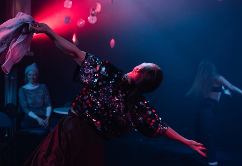 Kuvassa on tanssija selin vinossa asennossa molemmat kädet sivuilla ja vasemmassa kädessä on huivi. Tanssijalla on tumma glitterinen pusero.  Taustalla istuu tummuudessa tuolilla nainen harmaassa mekoss. Takana on punaista valoa.