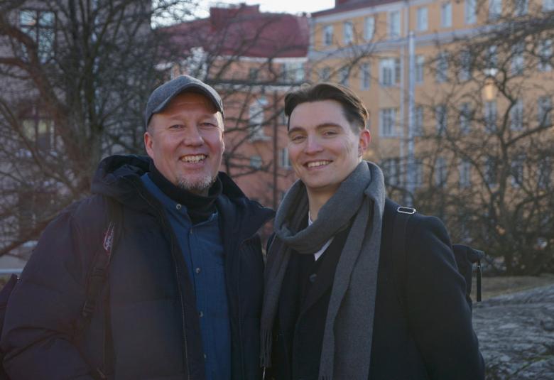 Kuvassa ovat Mikko Korsulainen vasemmalla tummassa takissa ja lippalakissa.  Oikealla on Rasmus Arikka mustassa takissa ja paljain päin.  Molemmat ovat puolivartalokuvassa ulkona ja taustalla näkyy paljaita lehtipuun runkoja ja oikealla on vaalea kerrustalo. 