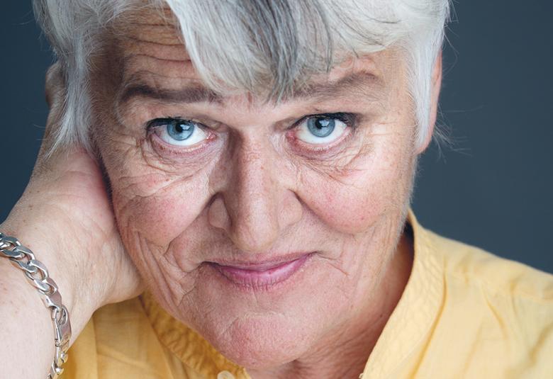 Kuvassa on Pirkkko Saisio kasvokuvassa ja hänellä on harmaat hiukset ja keltainen pusero. Hänellä on oikea käsi niskan takana.