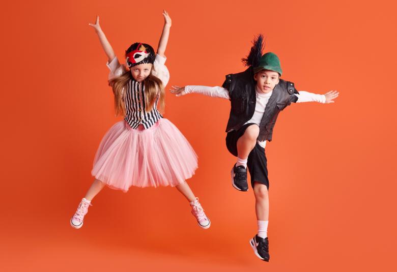 Kuvassa on vasemmalla hyppäämässä haara-perusasennossa tyttö,jolla on vaaleanpunainen hame. Oikealla poika tummassa liivissä ja polvihousuissa sekä sulkahatussa hyppyasennossa. Tausta on oranssi.