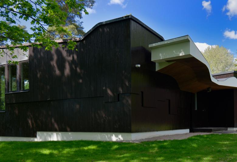 Kuvassa on Joonas Kokkosen talo ja se on tummaa puuta.  Vasemmalla on pystyt, korkeat ikkunat ja keskellä korkea, kaareva muoto ja oikealle suuntautuva siipi. Sen reunassa on neliömäinen ikkuna. 