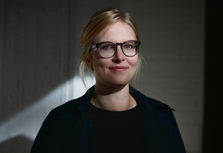 Kuvassa on keskellä Anna-Liisa Ahokumpu ja hän on puolivartalokuvassa. Hänellä on musta pusero päällään. Hänellä on isot silmälasit ja hiukset takana. Hänen takanaan on valojuova ja vasemmalla valoalue. Taustalla on tummaa.