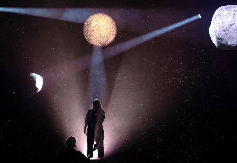  Kuvassa on tummalla pohjalla ylhäällä vasemmalla pieni kuu, josta tulee valoa ja keskellä keltainen kuu ja oikealla iso valkoinen kivimäinen tähti.  Keskellä on valoalue ja siinä on naishahmo seisomassa.