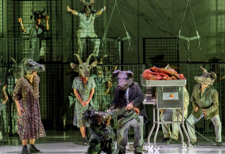 Kuvassa on taustalla vihreää verkkoseinää ja sen edessä on häkeissä eläinhahmoisia laulajia vihertävissä puvuissa. Etualalla on vasemmalla nainen lehmänpäällä ja tummassa mekossa. Etualalla on tummassa sikapäähineessä mies ja hän vetää tummassa lehmäpäähineesä olevaa hahmoa itseään päin.  Vieressä on tehine, jonka päällä on lihakimpaleita. Molemmin puolin on hahmot, joilla on vuohi- ja sikapäähineet.