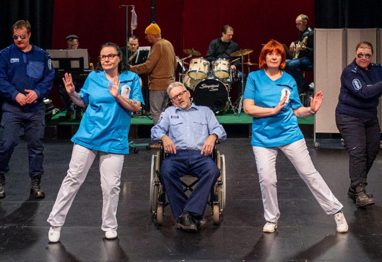 Kuvassa on kaksi sinisissä ja valkoisissa asuissa olevaa naishoitajaa ja heidän keskellään on poliisin puvussa ja pyörötuolissa. Molemmissa reunoissa on miespoliisit. Taustalla on viisi henkinen bändi.