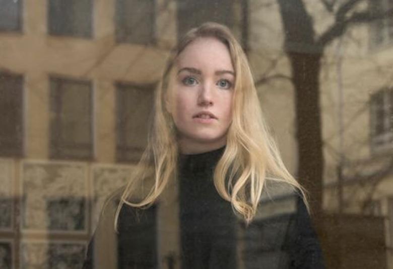 Kuvassa on Emilia Tanner puolivartalokuvassa. Hänellä on vaaleat pitkät hiukset ja musta poolopusero päällään. Taustalla on lasin takana ruskehtava kerrostalo.