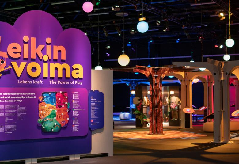 Kuvassa on tummahkoa tilaa ja katossa on pyöreitä valopalloja sekä tähtiä. Vasemmalla on pahviseinä, jossa on mm. isoin kirjaimin Leikin voima violetilla pohjalla. Taaempana näkyy ruskeita pylväitä.  