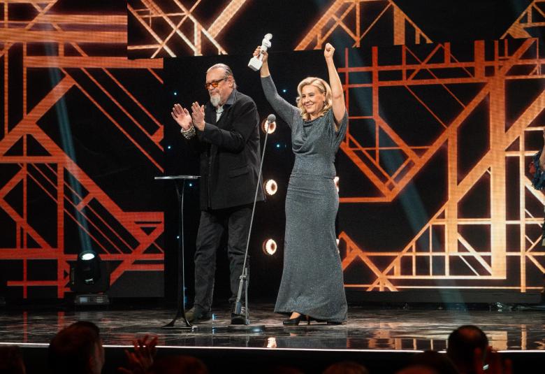 Kuvassa on vasemmalla Markus Selin tummassa puvussa ja taputtaa käsiään. Hänen oikealla puolellaan on Hanna Virolainen pitkässä harmaassa mekossa. Hänellä on kädet ylhäällä ja oikeassa kädessään palkinto.  Taustaalla on vinosti kuvioitua  poikkipuilla ja punertavaa väriä.