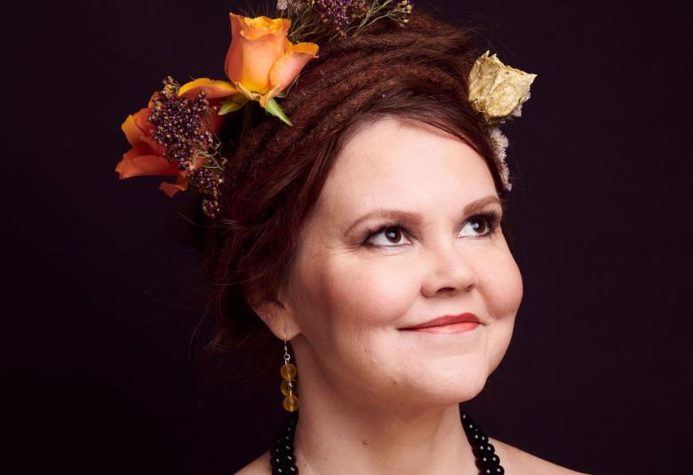 Kuvassa on Katja Ketun kasvot ja hänen päässään on iso, tumma nuttura.  Siinä on oranssin värisiä ruusuja koristeena. Kuvan tausta on tumma.