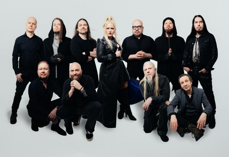Kuvassa on 13 muusikon ryhmä mustissa vaatteissa. He ovat kahdessa rivissä ja taaemmat seisovat. Tausta on vaalea.