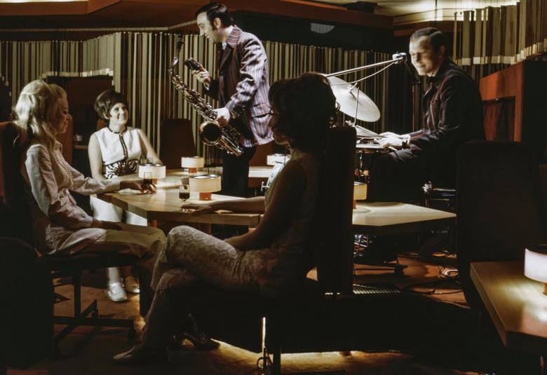 Kuvassa on pyöreän pöydän ääressä korkeaselkäisissä nojatuoleissa tummahkossa tilassa oikealla ja edessä katsomassa bändiä. Taustalla on saksofonisti ja oikealla rumpali soittamassa. 