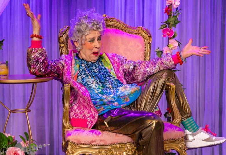 Kuvassa vanha nainen meikeissä ja värikkäissä vaatteissa istumassa kultareunaisessa nojatuolissa ja jalka käsinojalla. Taustalla on violettia kevytverhoa ja kukkia lattialla ja verjoissa. 