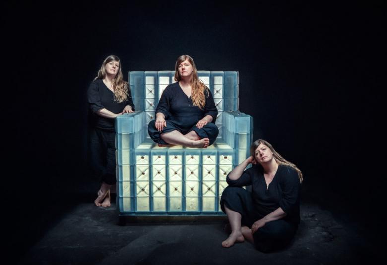 Kuvassa on iso lasinen vihertävän sininen nojatuoli ja edessä on vaaleita neliöitä. Marja Hepo-aho on mustassa puserossa ja pitkissä housuissa. Hepo-aho istuu tuolissa risti-istunnassa, tuolin edessä oikealla ja seisoo tuolin vasemmalla puolella.