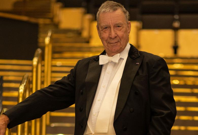 Kuvassa on Okko Kamu seisomassa postaikossa ja hän pitää oikealla kädellään metallisesta kaiteesta kiinni. Hänellä on musta frakki päällään ja kuva on puolivartalokuva. Kuvan sävy on kellertävä.