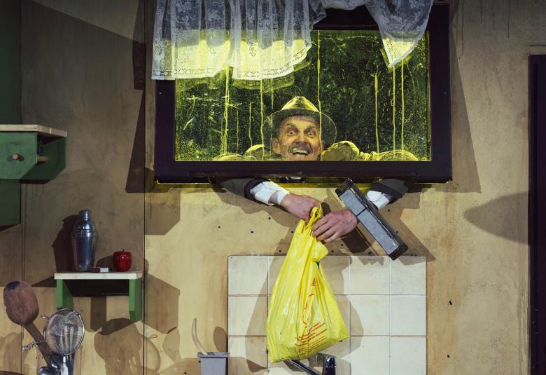  Kuvassa on huone, jossa on ikkuna ja siitä kurkistaa Marc Gassot.  Hän roikuttaa käsissään huoneen sisään keltaista pyyheliinaa. Huoneessa näkyy harmaata seinää ja ikkunan alla on valkoinen kaappi. 