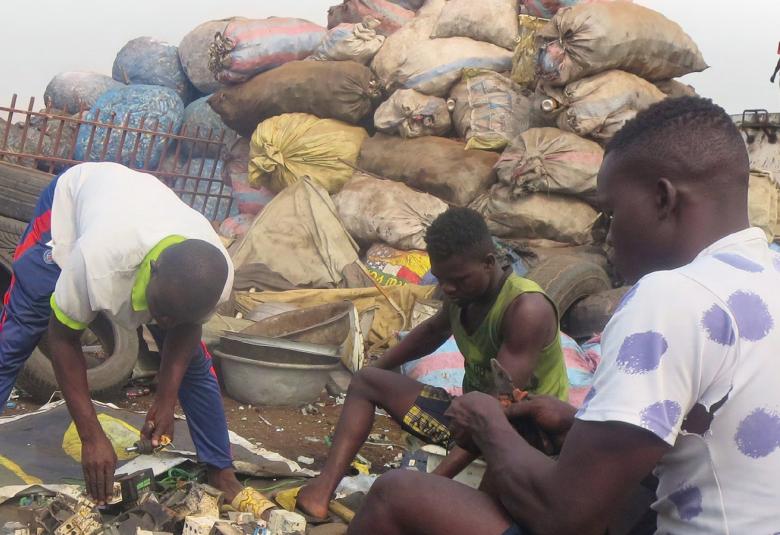 Kuvassa on kolme tummaihoista nuorta  miestä keräämässä roskia ja taustalla on vasemmalla renkaita. Takana on iso kasa säkkejä. Kuva on osa kirjan kannesta.