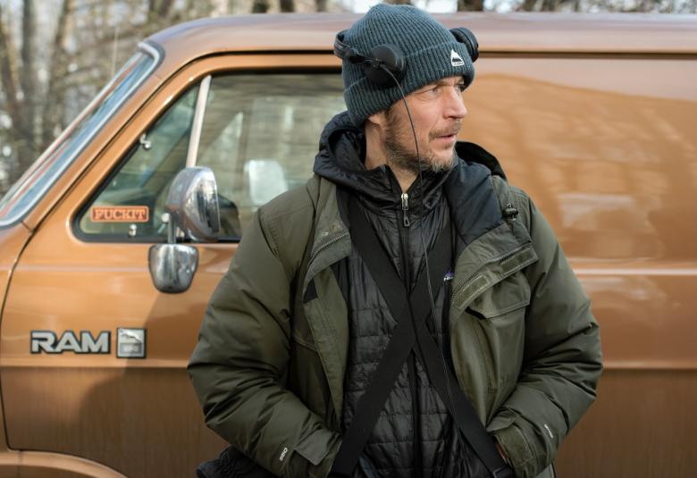 Kuvassa on Aleksi Salmenperä seisomassa sinapin keltaisen pakettiauton edessä. Hänellä on tumma pusakka päällään ja pipo päässaan. Katse on käännettetynä oikealle ja kädet taskuissa.