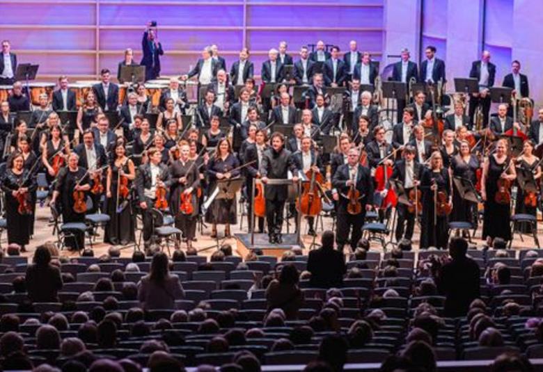 Kuvassa on Tampereen Filharmonia seisomassa lavalla soittimet kädessään ja mustissa puvuissa. Etualalla on penkkiriveissä yleisöä ja osa seisomassa.