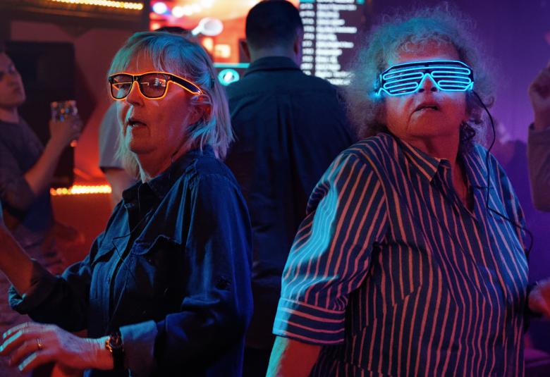 Kuvassa ovat vasemmalla Leena Uotila ja vasemmalla Saara Pakkasvirta diskossa tummassa valaistuksessa.  He ovat tanssimassa puolivartalokuvassa ja heillä on kuvioidut valolasit päässään.