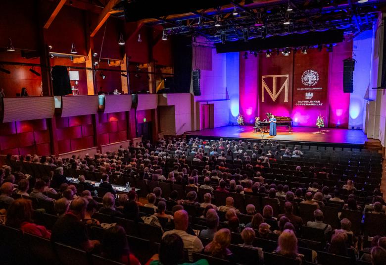 Kuvassa on Savonlinna-sali melko pimennettynä ja yleisöä näkyy takaapäin alaspäin laskeutuvassa katsomassa. Edessä on pinkki-violeteilla kankailla koristeltu näyttämö, jossa on sinisessä pikässä mekossa esiintyvä naislaulaja. Säestäjä on takana flyygelin ääressä.