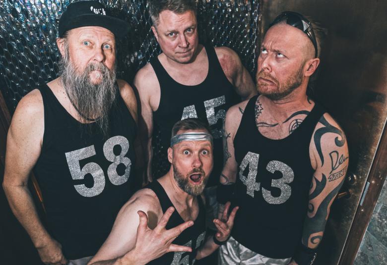  Kuvassa on neljä Wöyh!in miesjäsentä kuvattuna ylhäältä päin. He seisovat tiiviisti ryhmänä ja heillä on hihattomat musta paidat sekä yhtä lukuunottamatta tatuointeja. Yksi on alempana ja hän haristaa oikean käden sormiaan.