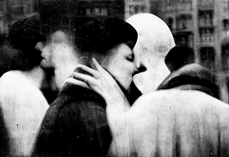 Kuvassa on keskellä vanhempi nainen mustassa takissa ja hatussa sekä vieressä toinen nainen selin. Kuvassa näkyy valkoisia ja mustia heijastumia.  Taustalla näkyy himmeänä kerrostalo. Kuva on musta-valkoinen.