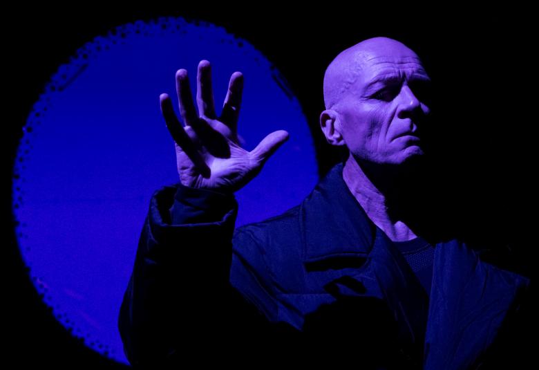 Kuvassa on Pentti Korhonen puolivartalokuvassa mustassa takissa ja hänellä on oikea käsi pystyssä.  Taustalla on sininen iso kuu ja muu osa taustasta on musta.