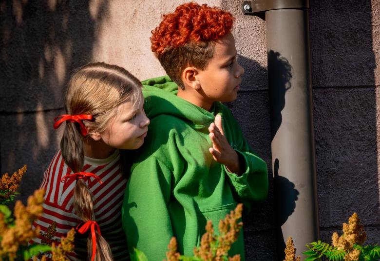 Kuvassa ovat kurkksimassa talon nurkalta etummaisena Aleksi Johansson ja Alma Järvensivu.  Johanssonilla on punaista kiharaisissa hiuksissaan ja vihreä huppari. Järvensivulla on saparot ja tummaraidallinen pusero.