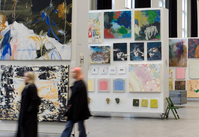 Kuvassa on taustalla isolla ripustusalustalla useita taideteoksia ja kuvassa näkyy niitä kolmessa rivissä.  Edessä kulkee kaksi ihmistä himmennettyinä ja heillä on mustat takit päällään.