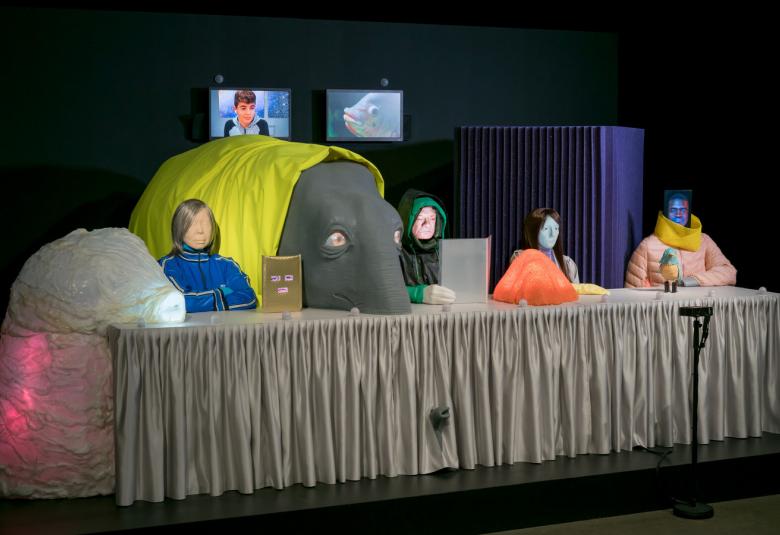 Kuvassa vaalean ruskean kankaan päällä rintakuvallisia erivärisiä nukkeja neljä ja keskellä on keltaisella kankaalla päällystetty norsin pää. Tausta on tumma.