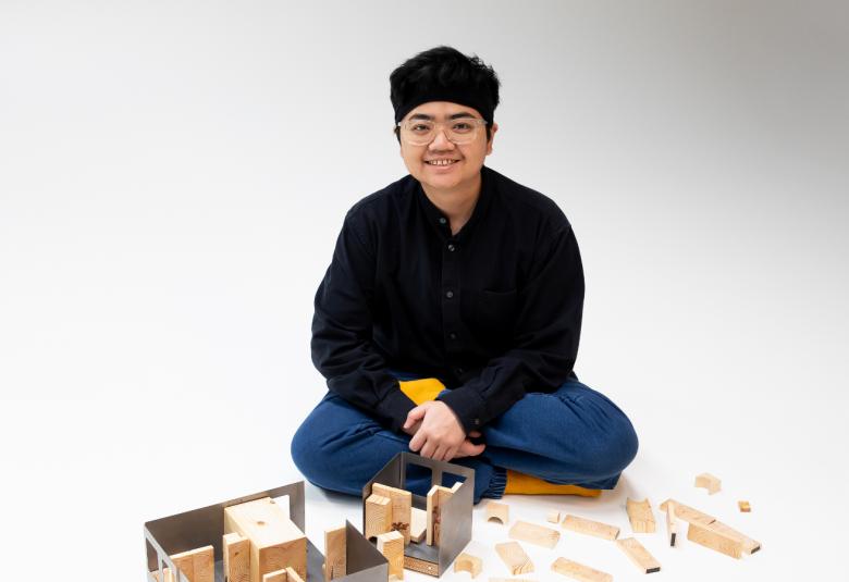 Kuvassa on Nhung Pham risti-istunnossa mustassa asussa ja hatussa istumassa puusta tehdyn talon pienoismallin edessä ja siinä on erikseen pienoismallin osia. Tausta on vaalea.