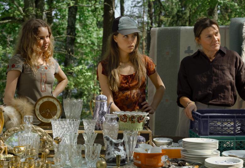 Kuvassa ovat täynnä laseja ja lautasia olevan pöydän takana seisomassa Ada Pesso vasemmalla, Aamu Milanoff ja Pirjo Lonka astia kori käsissään. Takana on lehtimetsää ja mattoja narulla.