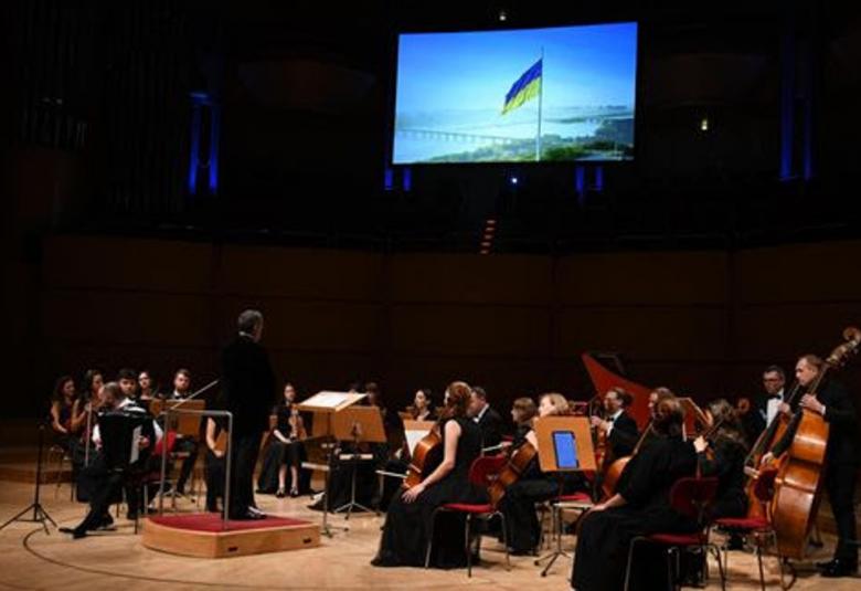 Kuvassa on sivulta päin kuvattuna Four Seasons -orkesteri ja vasemmalla keskellä seisoo orkesterikorokkeella. Tausta on tumma ja siinä on heijastettu Ukrainan lippu liehumassa ja taustalla on maisemassa siltoja sekä merta.