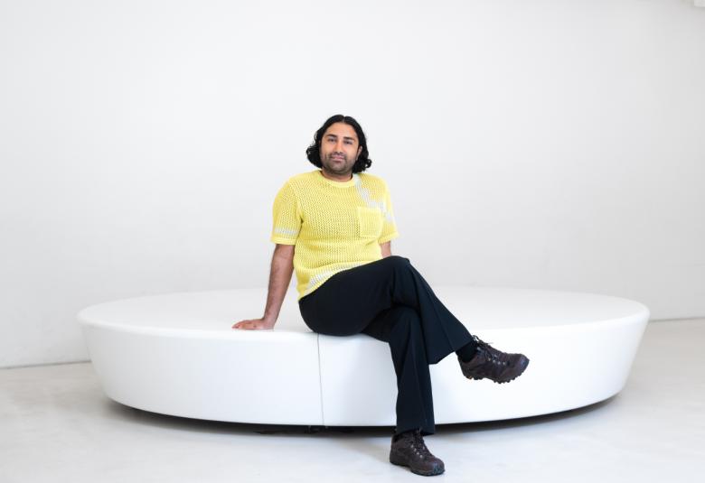 Kuvassa on ison, valkoisen pyöreän muotisen istuimen päällä istumassa Harold Hejazi. Hänellä on keltainen t-paita ja mustat housut. Tausta on vaalea.